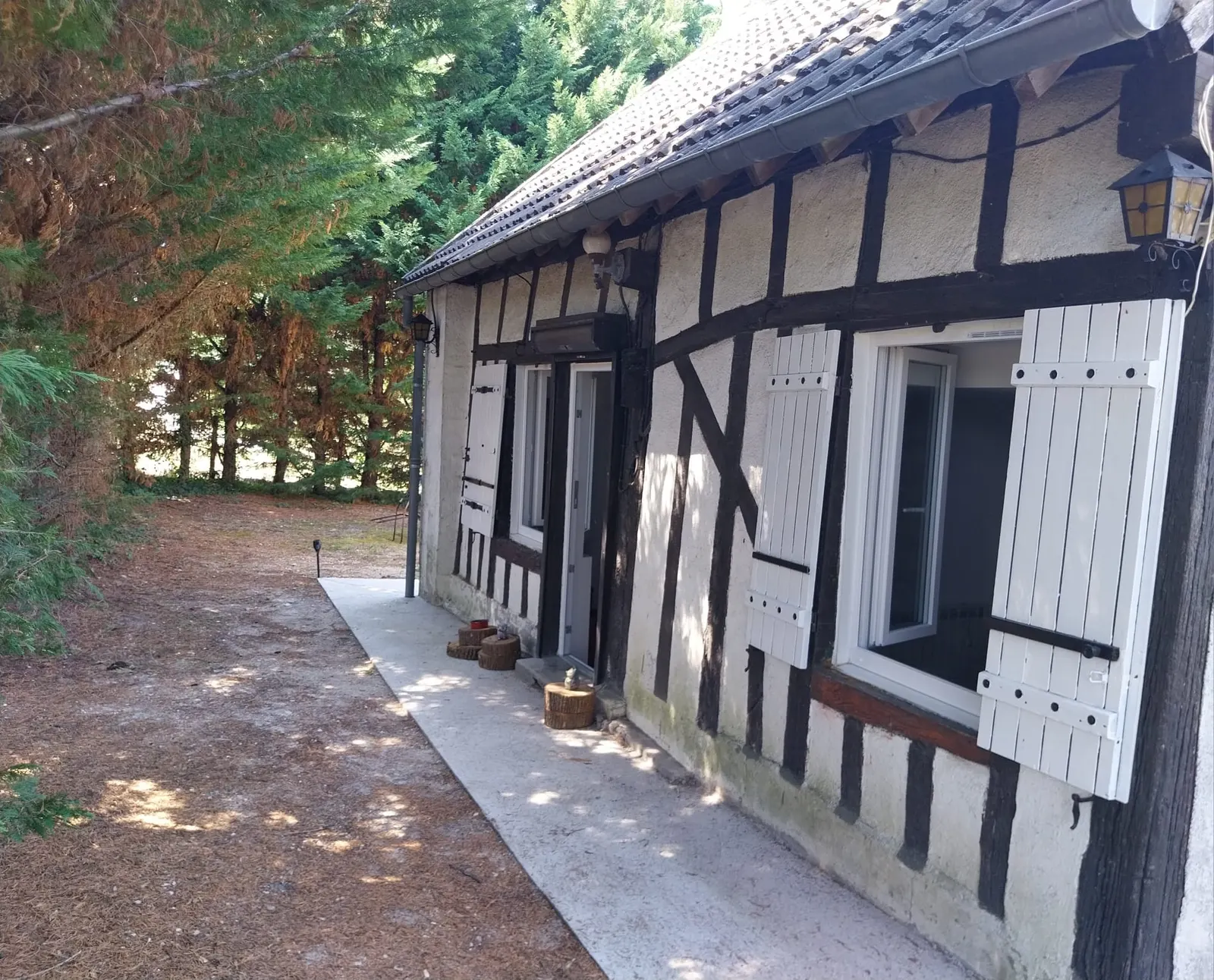 Le Répit : la façade à colombages de la maison, volets blancs ouverts sous les grands conifères