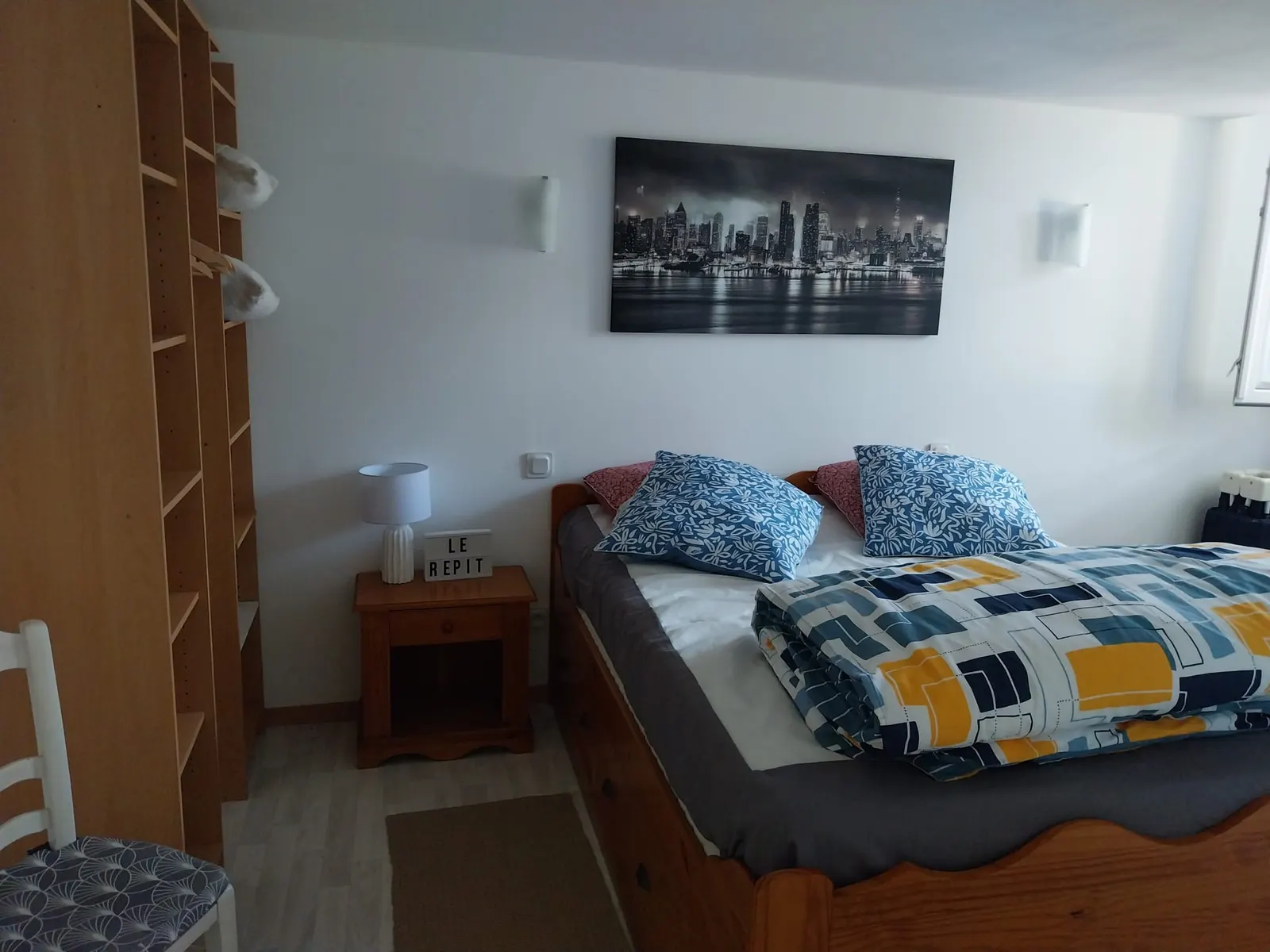 La chambre du Répit : lit double, coussins bleus et petit panneau lumineux « Le Répit » sur la table de chevet
