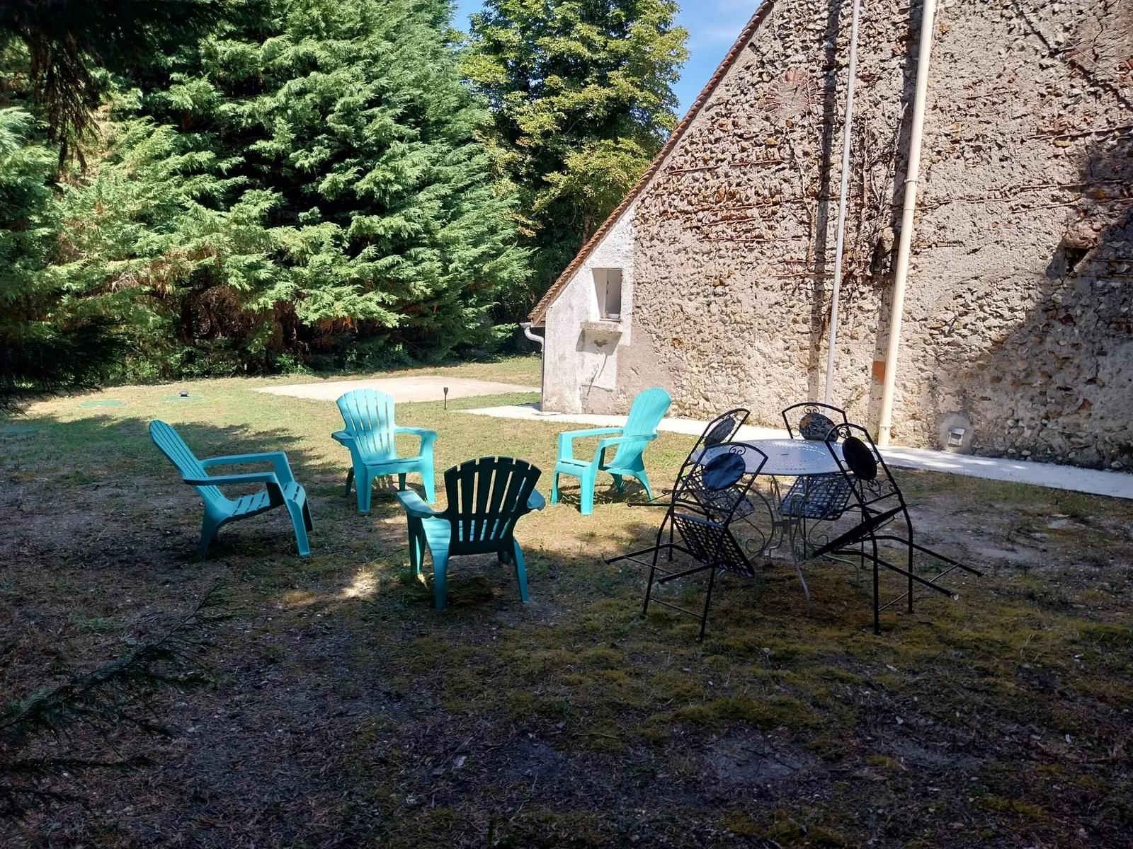 Fauteuils de jardin turquoise et salon en fer forgé sur la pelouse ombragée du Répit, au pied du pignon en pierre